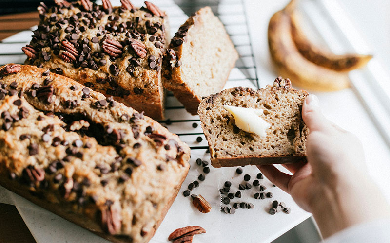 Loaves of baked bread on counter, hand holding slice with butter